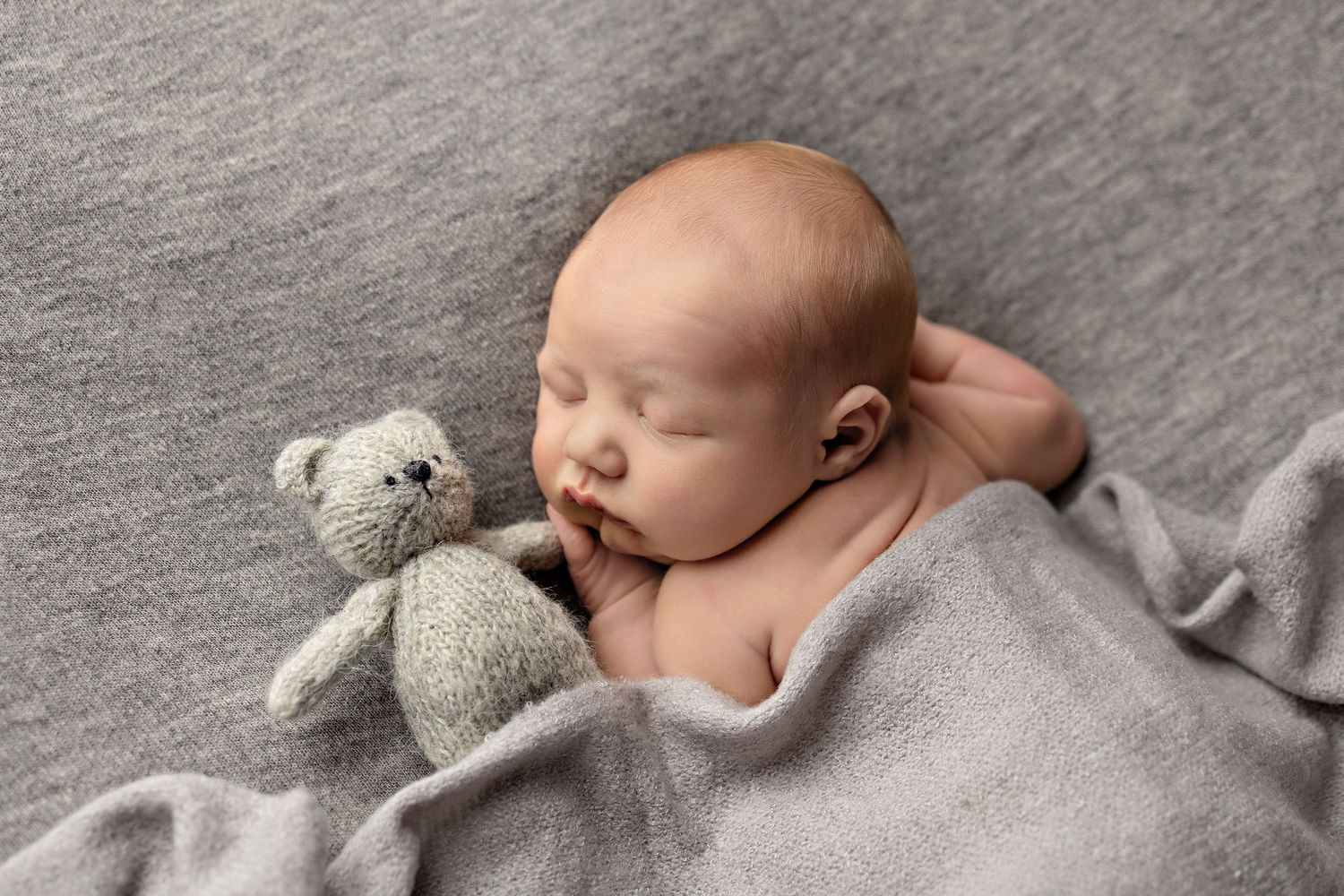 Peaceful newborn baby sleeping on gray blanket with a small knitted teddy bear, soft and serene nursery photography.
