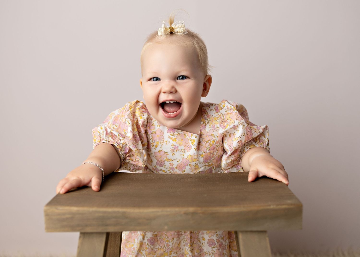 Happy baby girl in floral dress laughing in wooden high chair against neutral background.