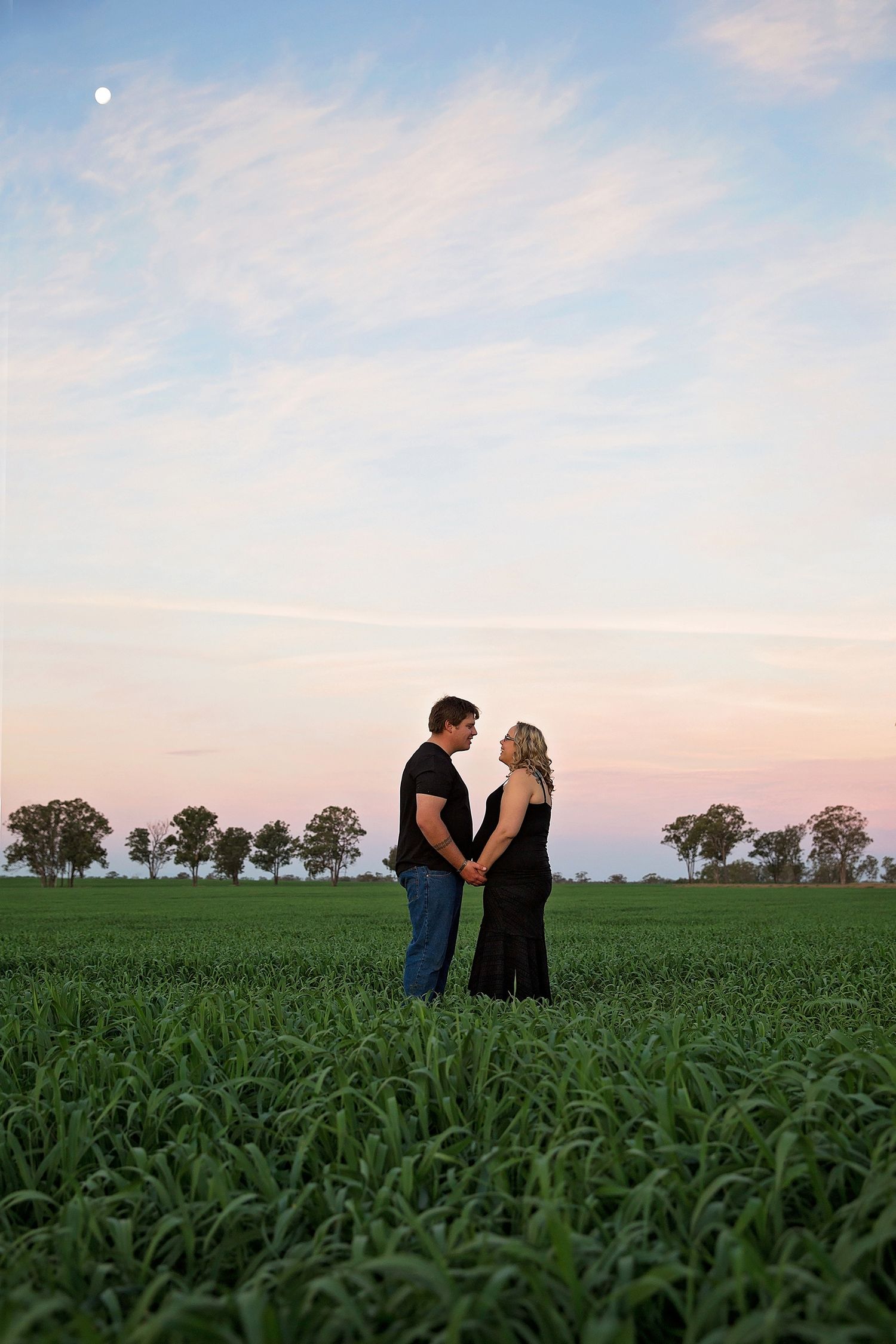Couple embraces in a vast green cornfield under soft evening sky.