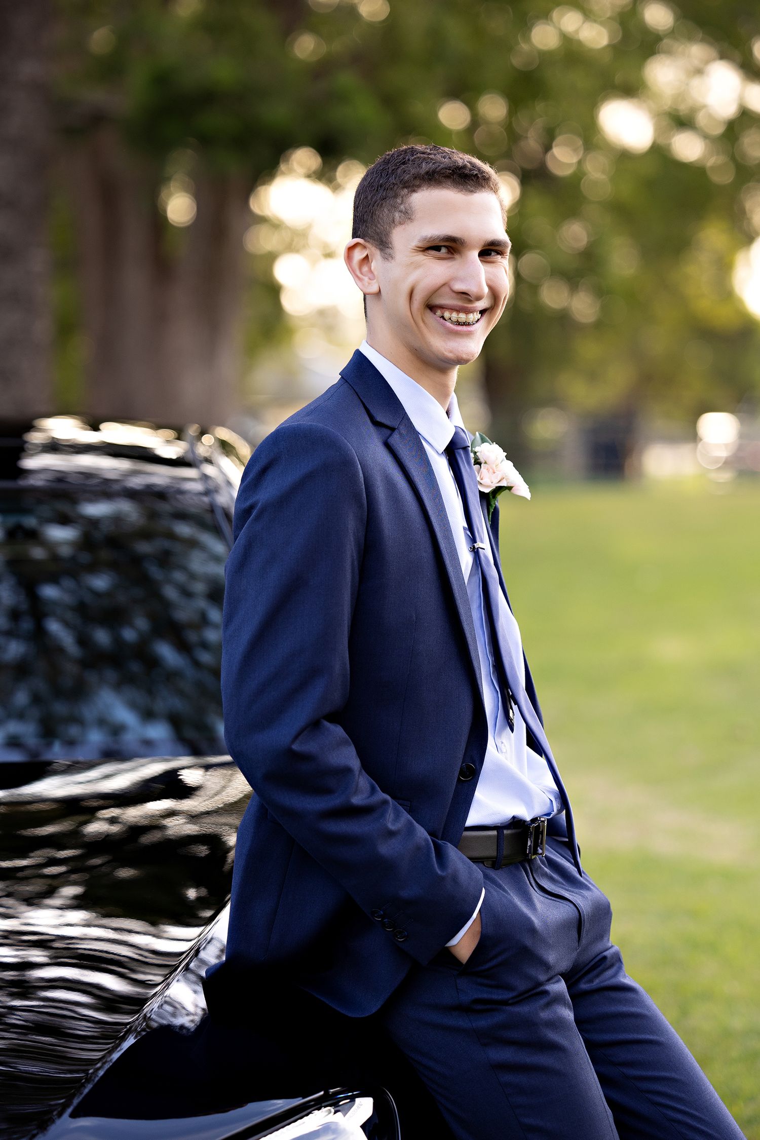 Person in navy suit smiles while leaning against black car outdoors.Toowoomba Photographer Toowoomba Newborn Photographer