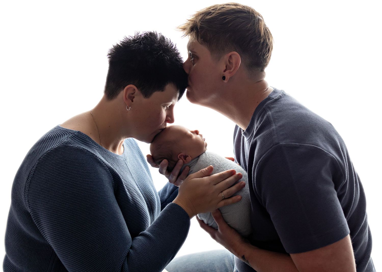 Two parents share a tender moment kissing over their sleeping newborn baby held gently between them.