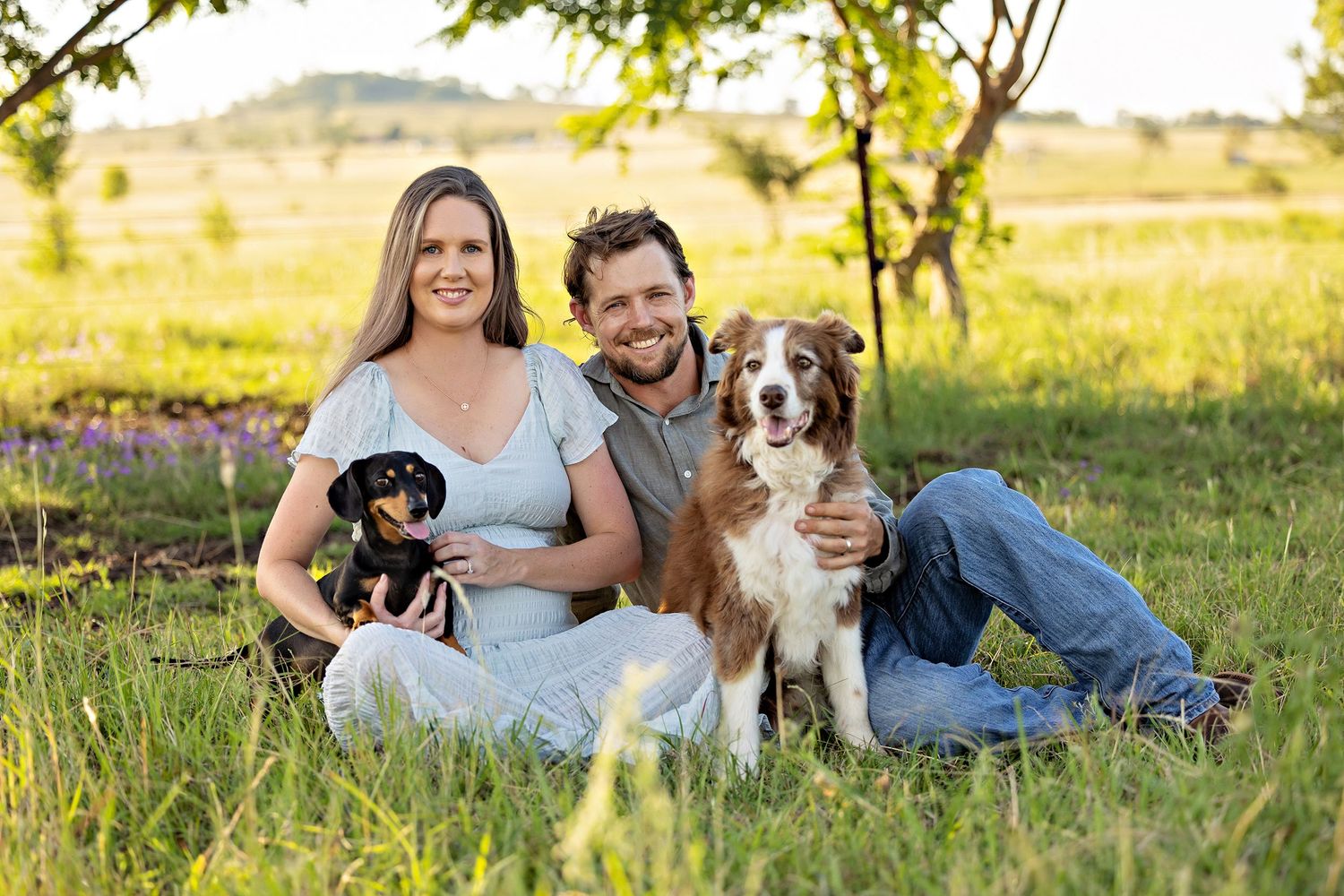 A couple relaxes on grass with their Australian Shepherd dog during a golden hour photoshoot.