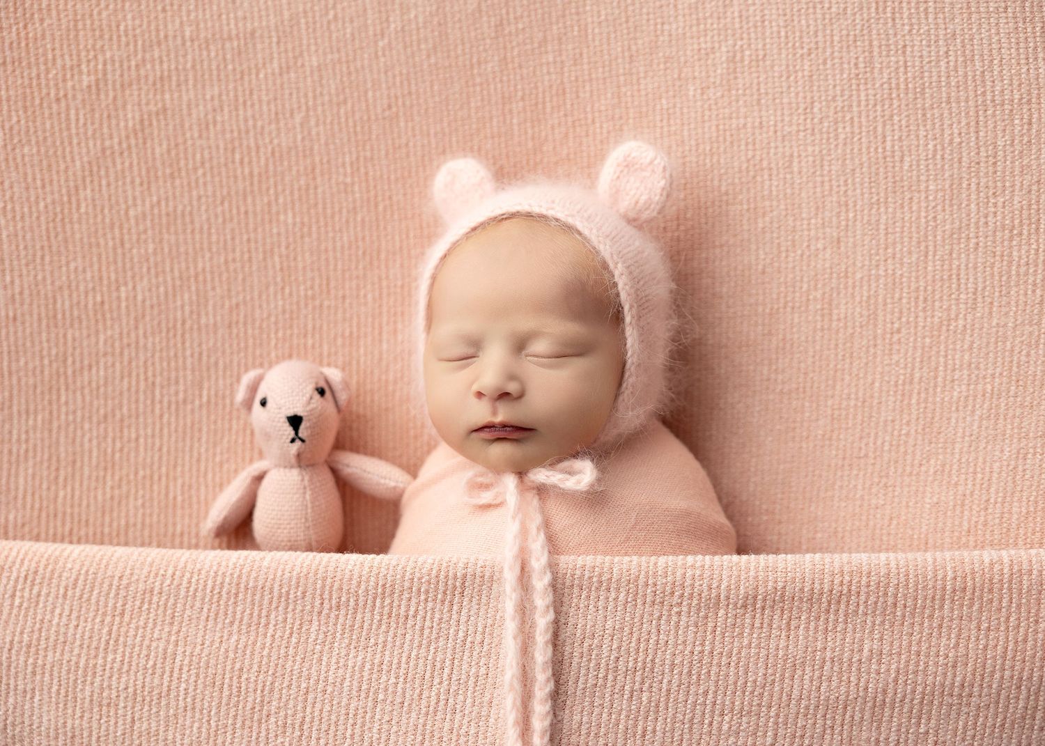 Sleeping newborn baby in soft pink tones with white teddy bear companion creating peaceful nursery scene.