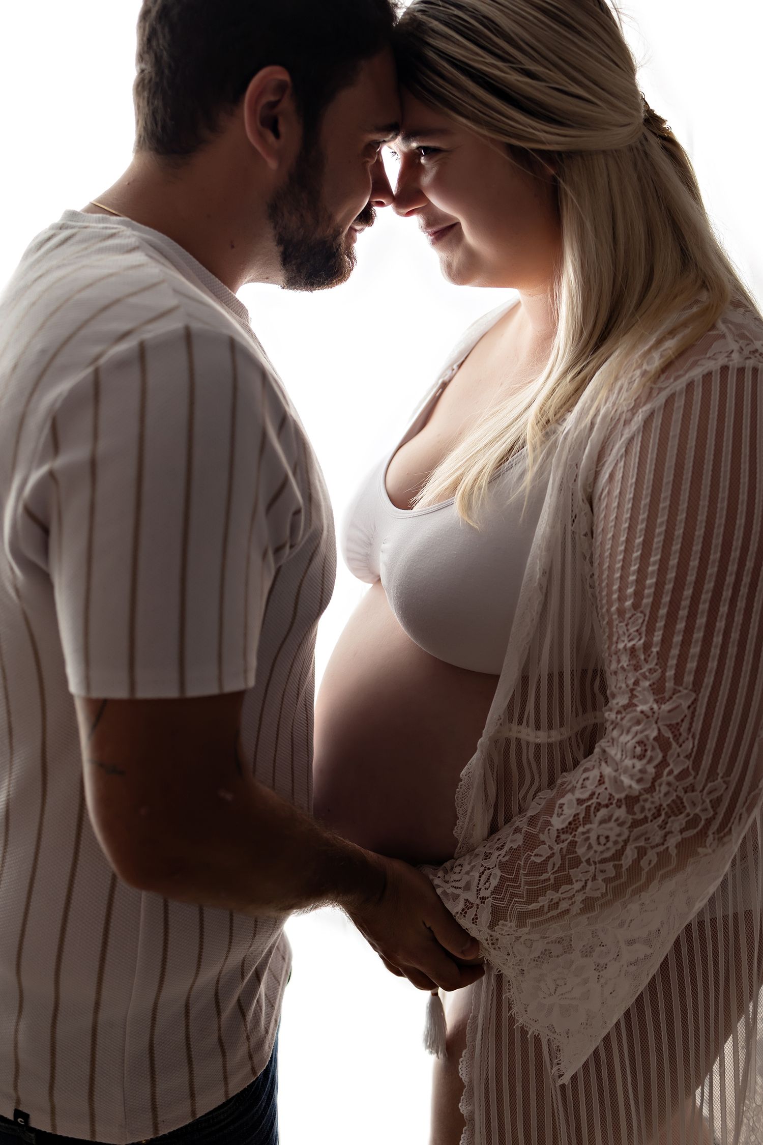 Couple sharing an intimate moment, pregnant woman in white lace robe holding hands with bearded man in striped shirt.