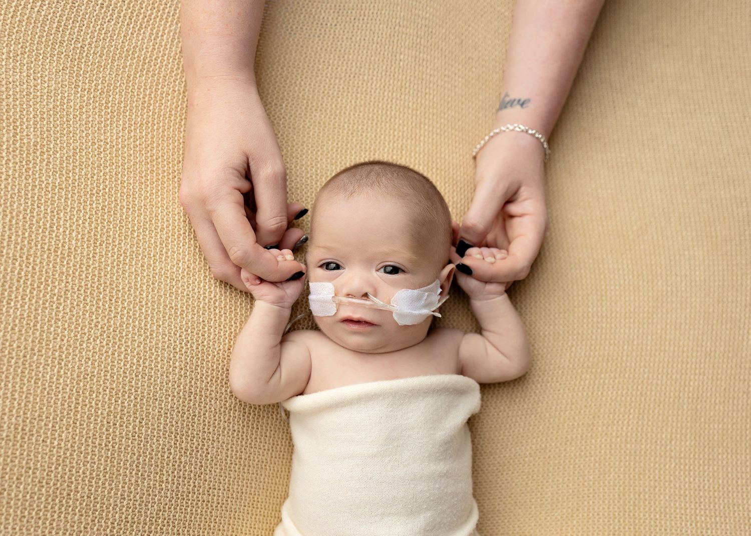 A small infant wearing a nasal cannula lies on beige fabric while hands gently touch their head.Toowoomba Photographer