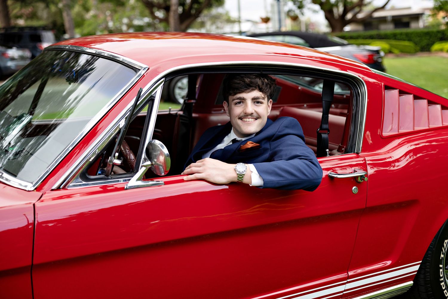 Young man in suit smiling from red classic Ford Mustang fastback window at outdoor car show.