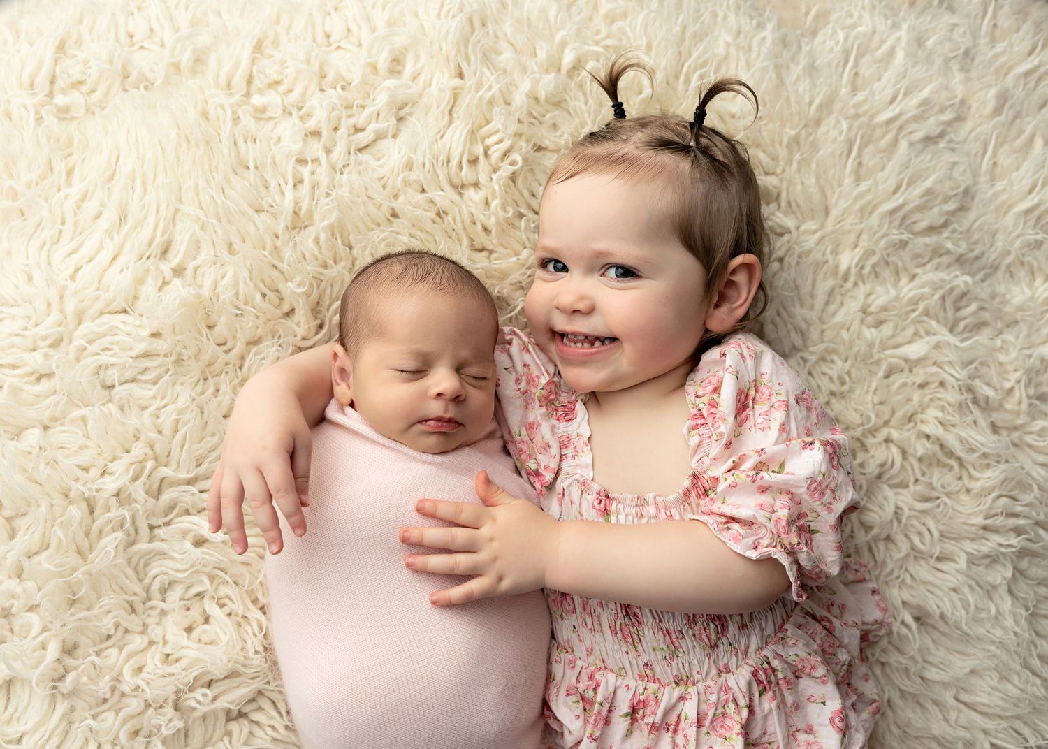 Two young children on cream fur rug, older sibling embracing sleeping newborn.