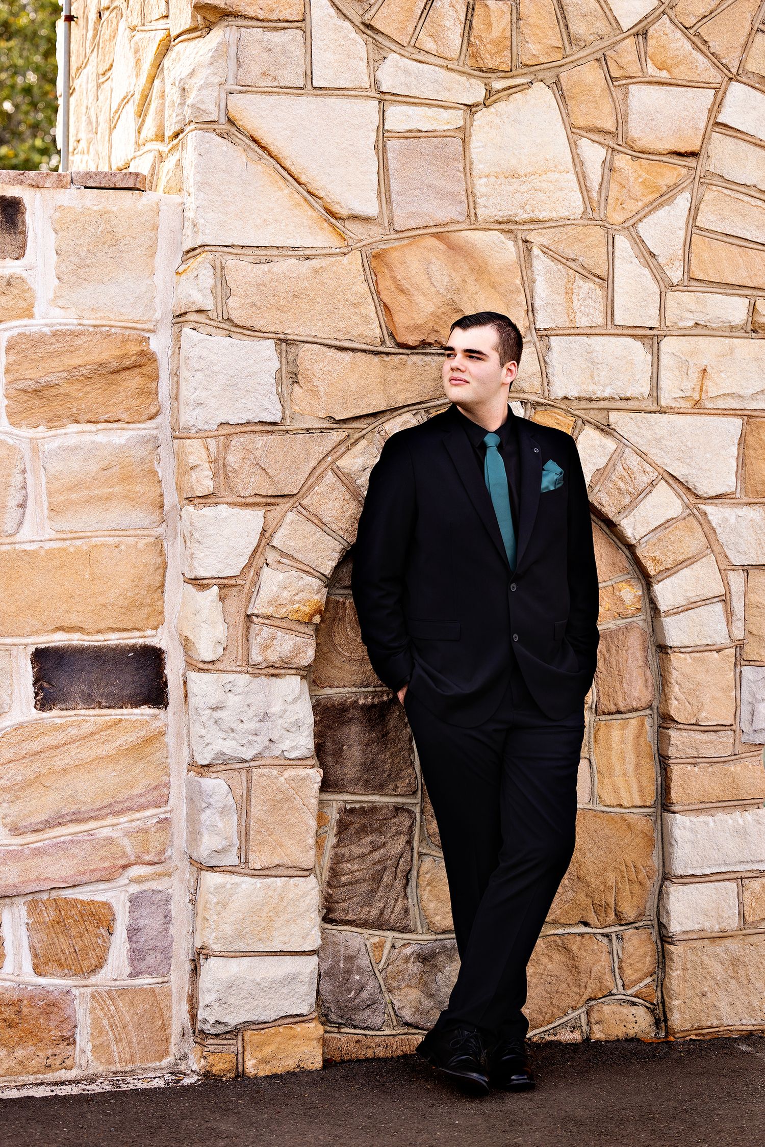 Person in black suit and teal tie poses against decorative stone wall architecture.Toowoomba Photographer Toowoomba Newborn Photographer