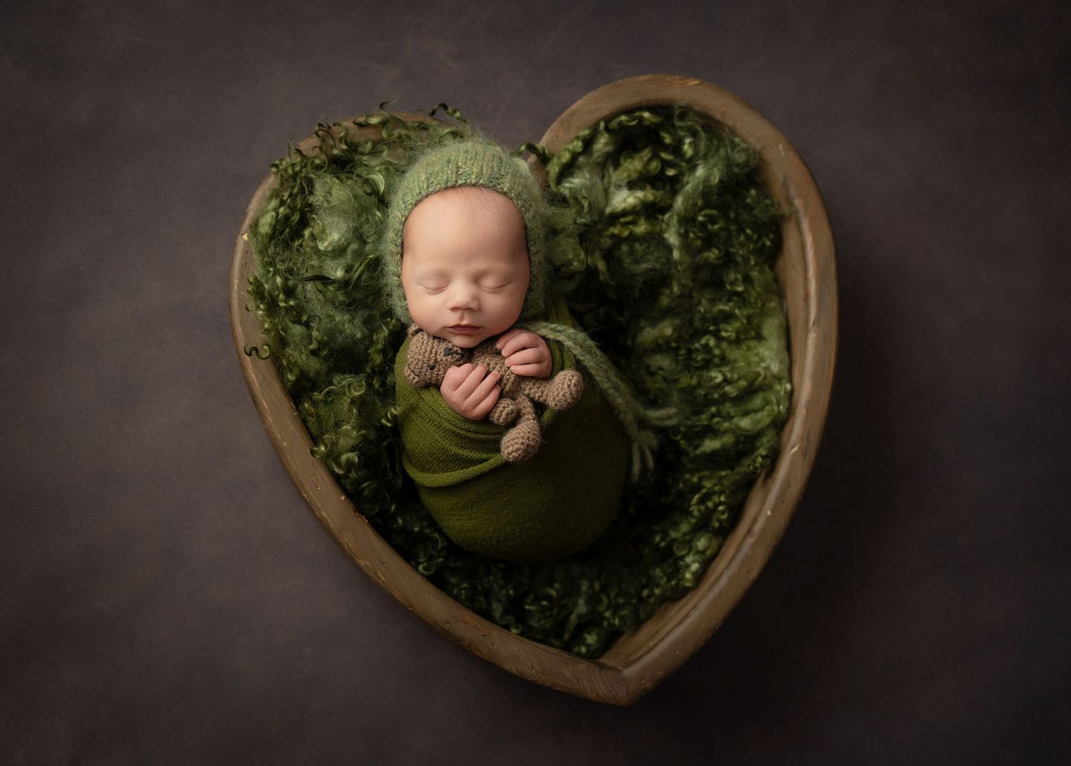 Sleeping newborn nestled in heart-shaped wooden bowl with green foliage.