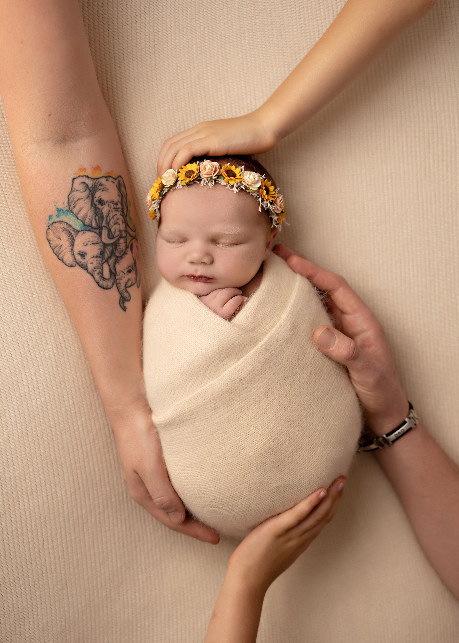Hands with a butterfly tattoo gently cradle a sleeping newborn wrapped in beige fabric against a neutral backdrop.