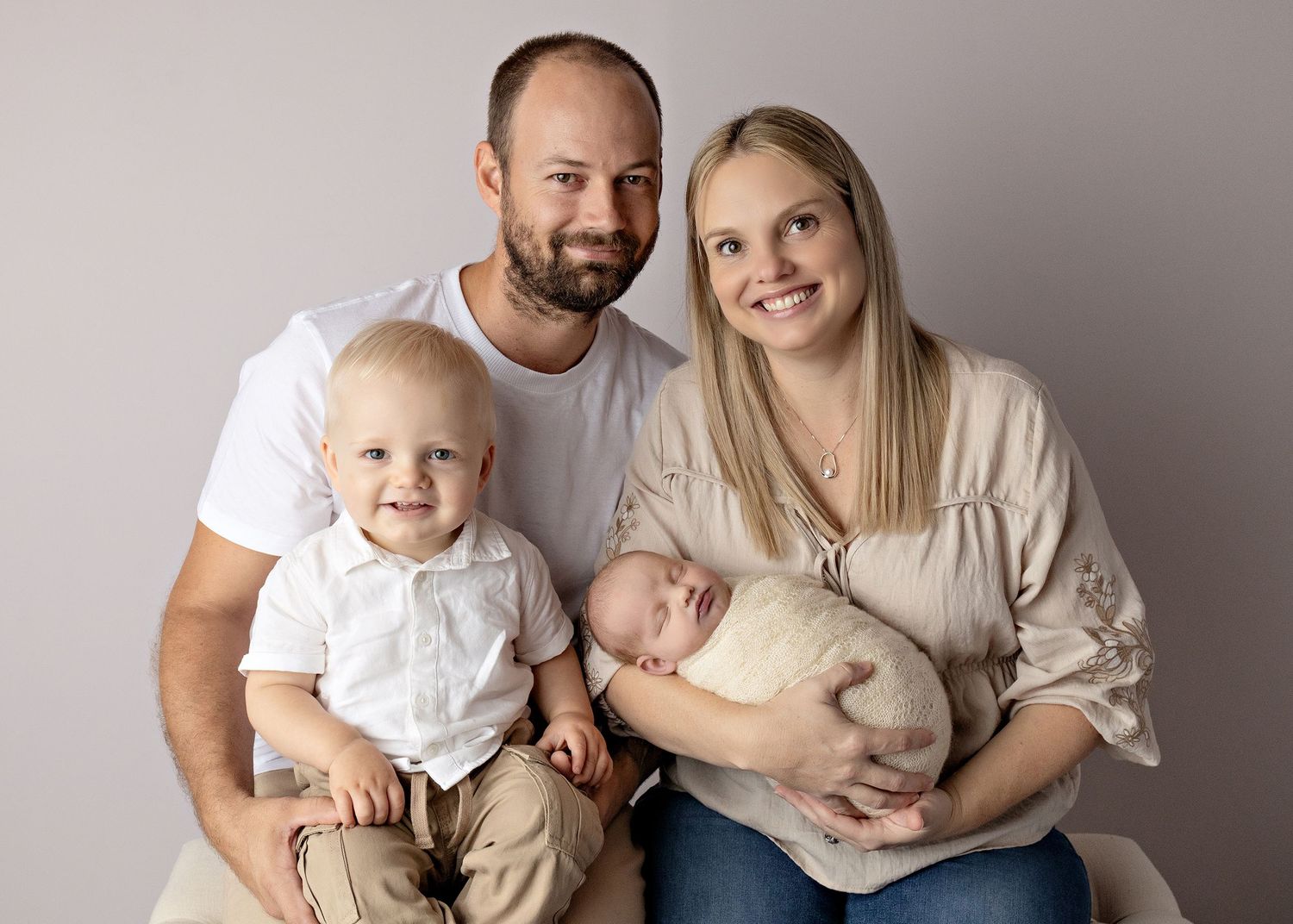 Family portrait of parents with toddler son and newborn baby posed together against a neutral gray background.