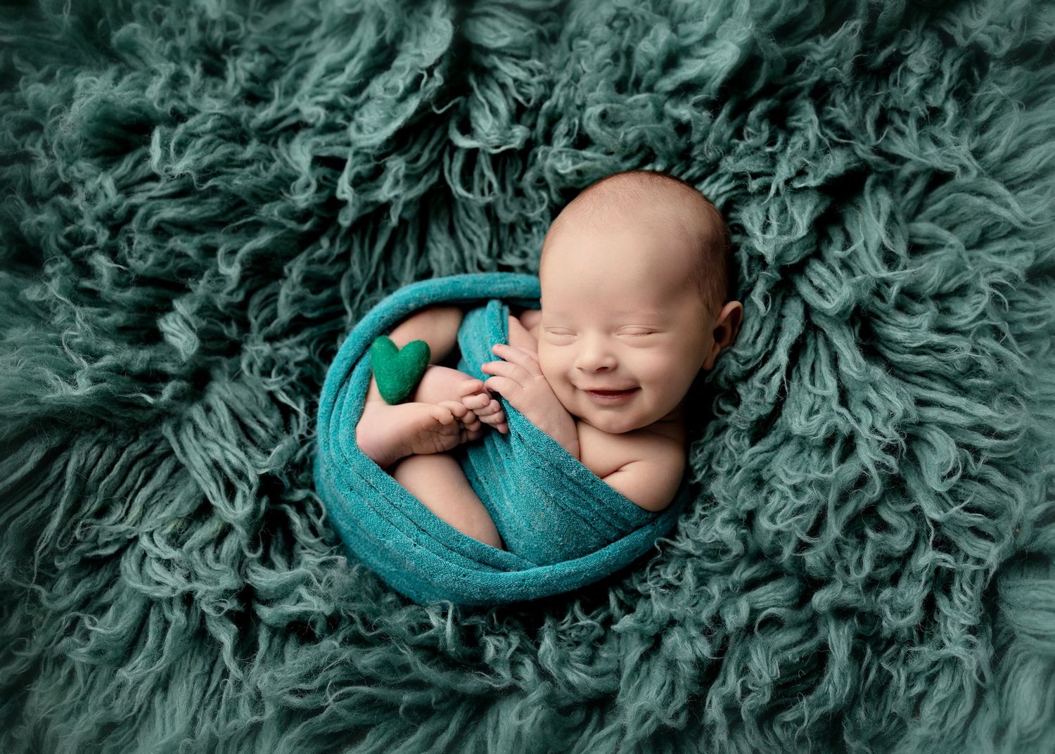 Smiling newborn baby wrapped in teal swaddled in a bowl on a teal fluffy blanket.