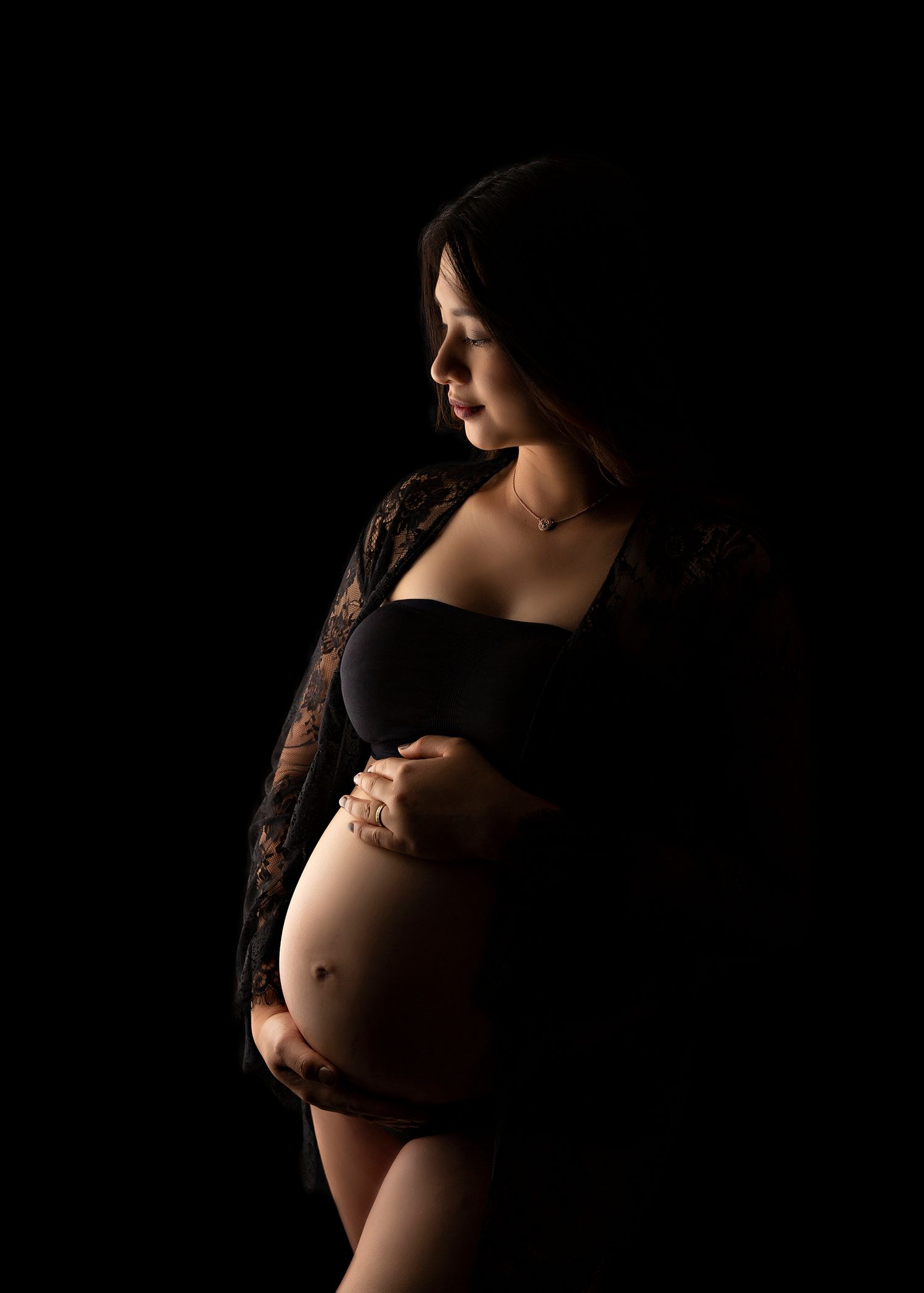 Dramatic maternity portrait with black lace and dark background highlighting pregnant silhouette.