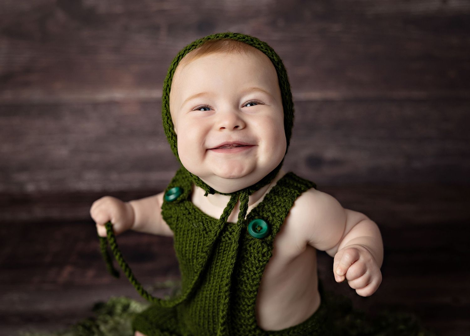 A baby wearing a green knitted vest and bonnet smiles joyfully against a dark wooden background.Toowoomba Photographer