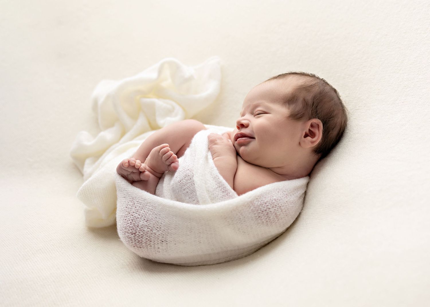 Peaceful newborn baby sleeping wrapped in a soft white swaddle blanket on a cream background.
