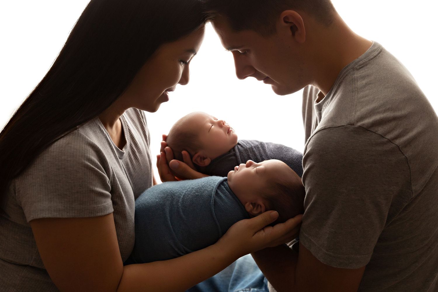 Parents sharing tender moment with newborn twins in backlit setting.