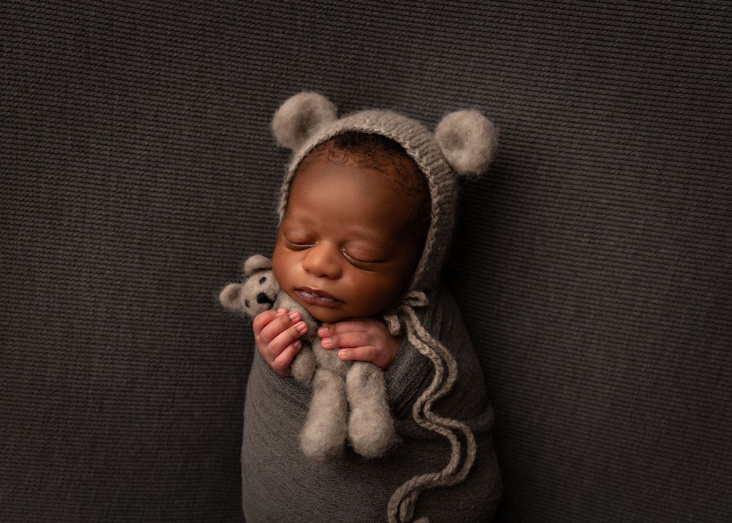Sleeping baby wearing gray knitted bear bonnet and wrapped in cozy gray swaddle on dark background.Toowoomba Photographer