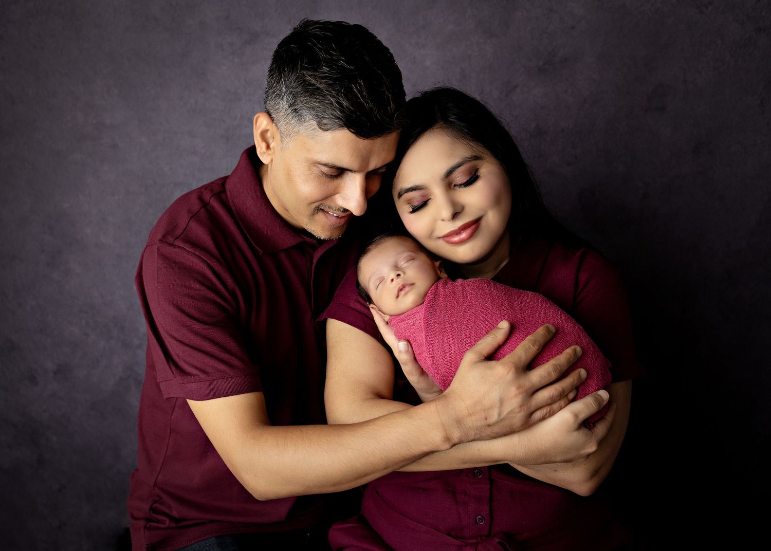 Parents in matching burgundy outfits tenderly hold their sleeping newborn wrapped in pink against a gray studio backdrop.