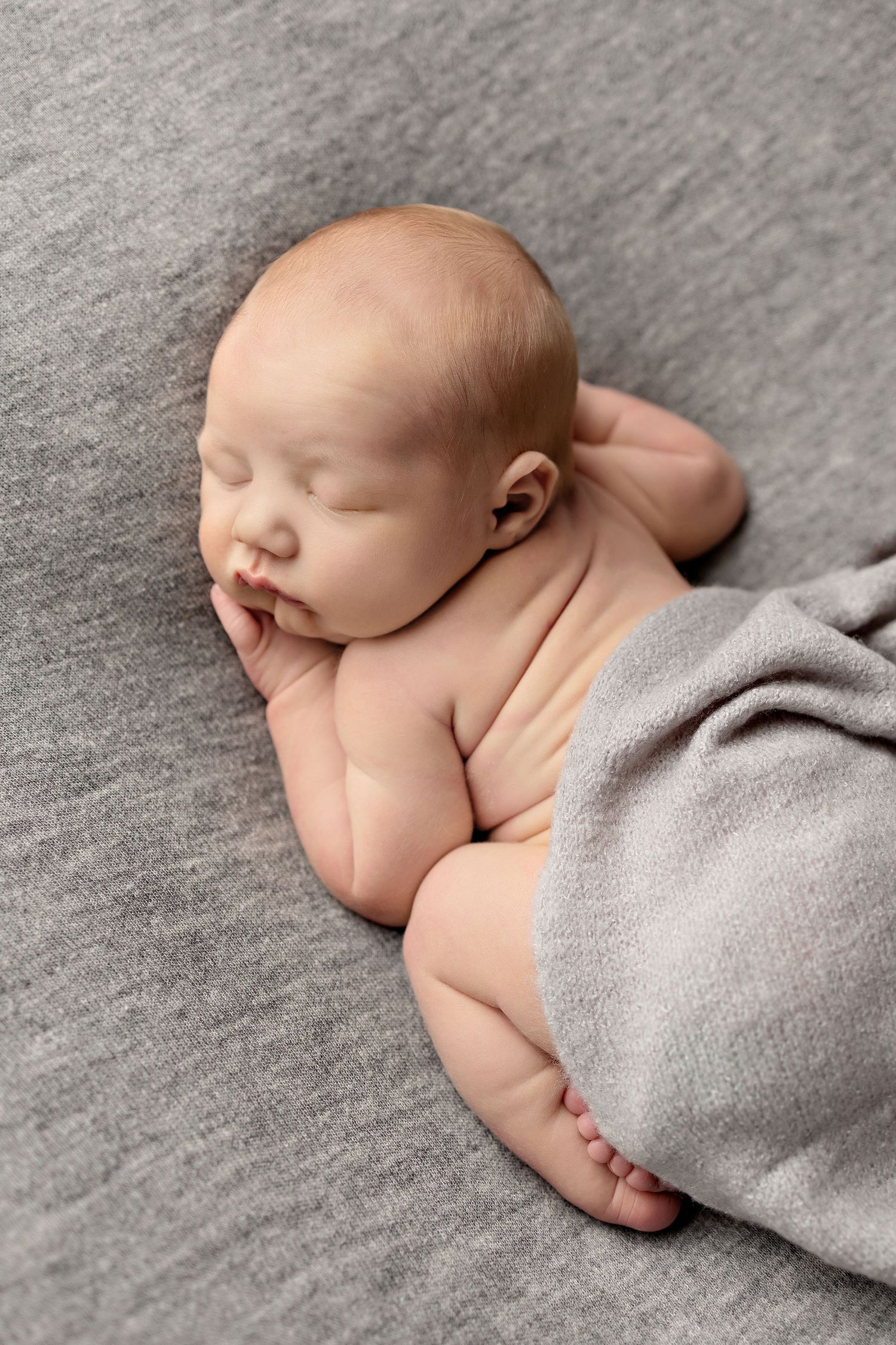 Peaceful newborn baby sleeping curled up on gray fabric, partially covered with a soft gray blanket.