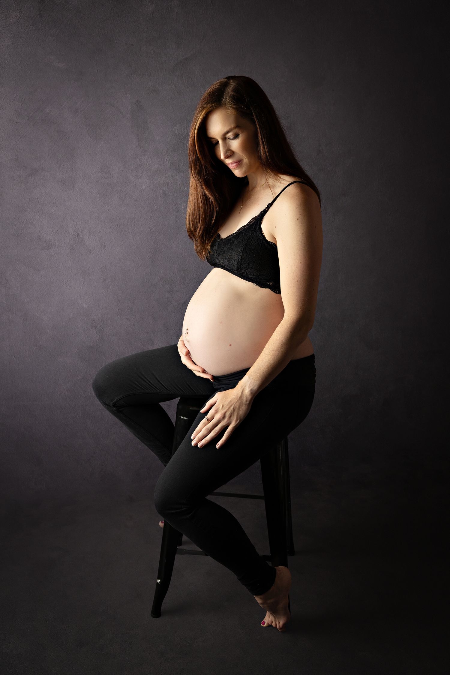 Artistic maternity portrait of woman in black outfit seated on stool against dark background.