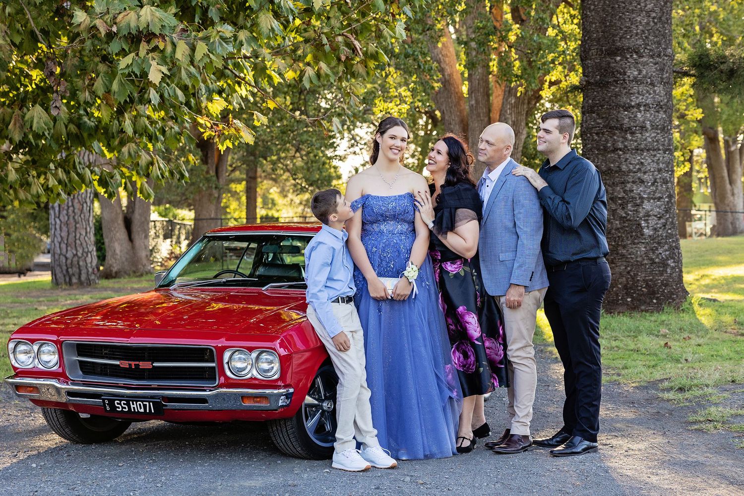 Family posing beside a classic red SS Holden muscle car outdoors for a formal occasion.