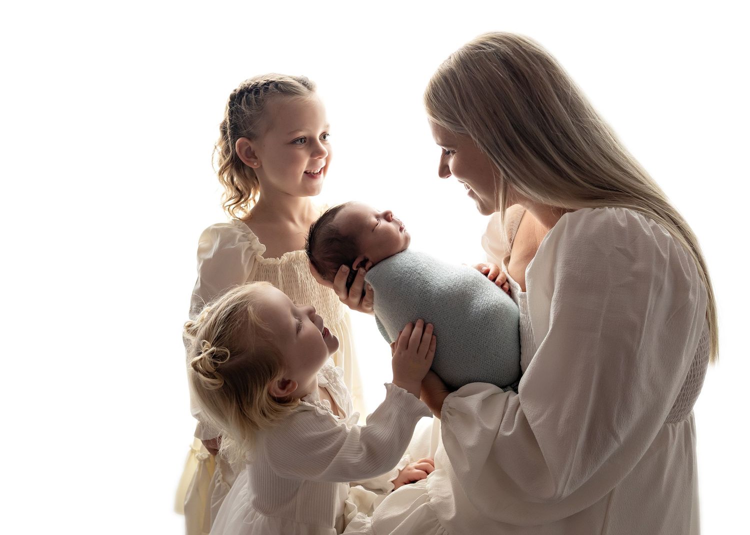 A mother in white holds a swaddled newborn while two young daughters in cream dresses look on, white background.