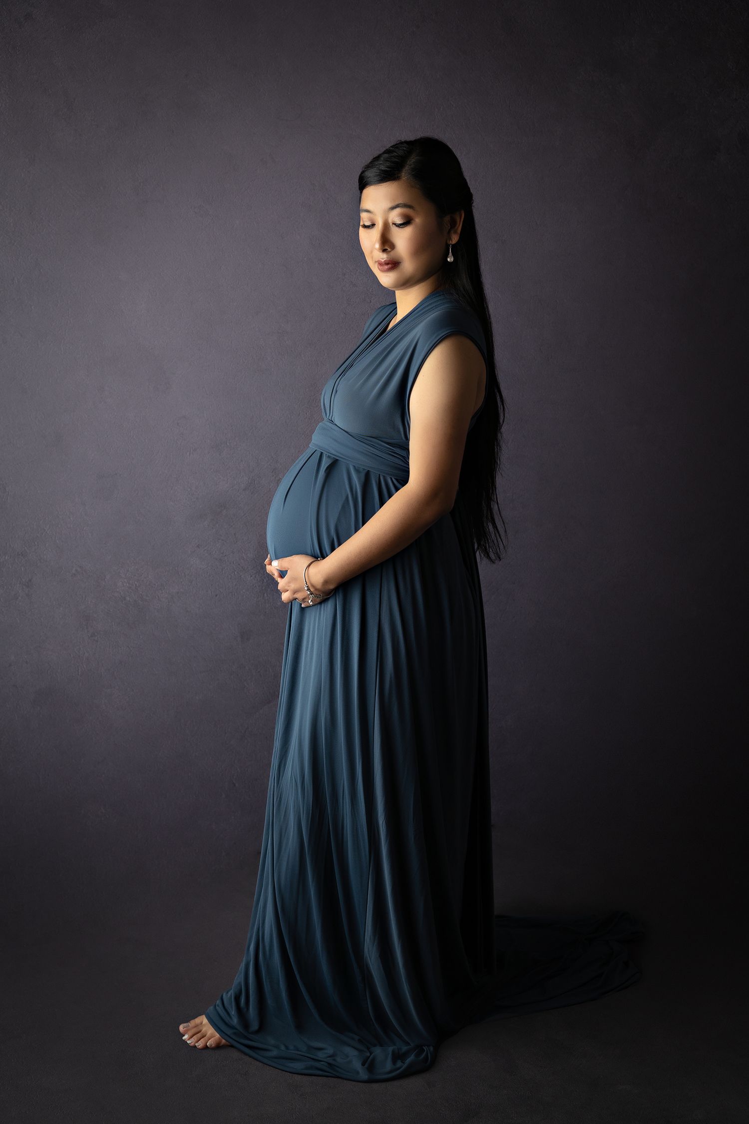 Elegant maternity portrait in flowing blue dress against dark background.