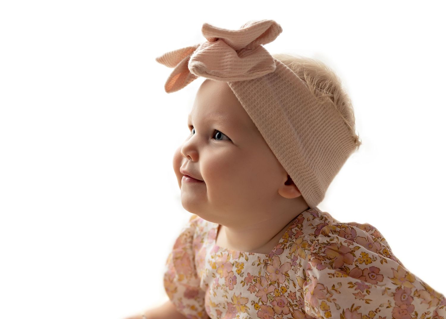 Smiling baby wearing a pink floral outfit and beige bow headband against a white background.