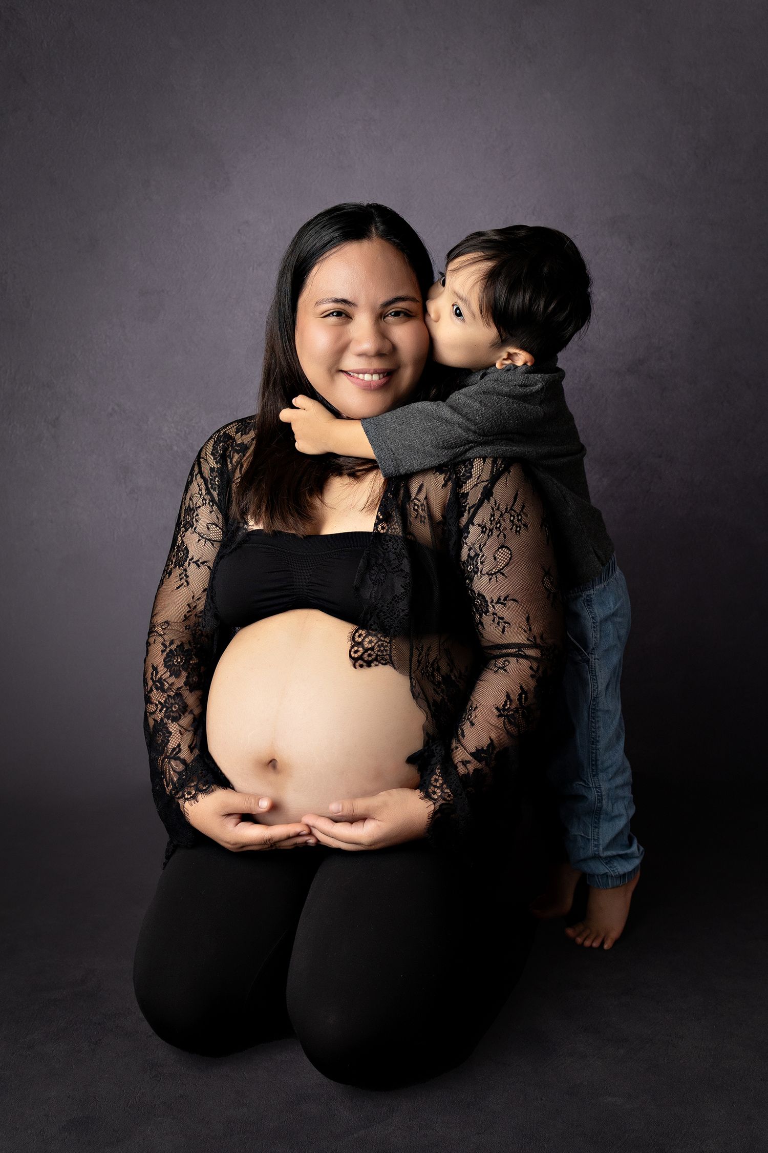 Pregnant woman in black lace outfit kneeling while young boy hugs and kisses her cheek in studio maternity photo.