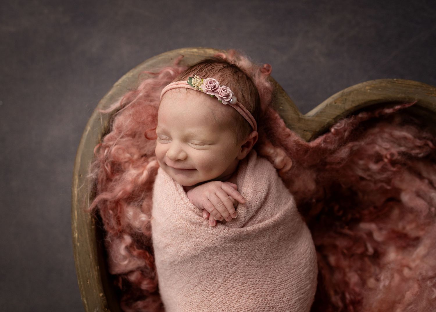 Smiling newborn baby wrapped in pink swaddle sleeping in heart-shaped wooden bowl with floral headband.