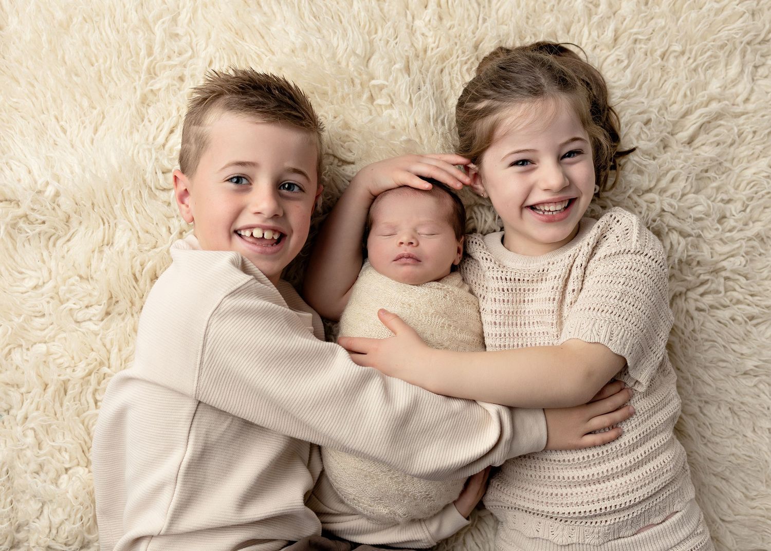 Two smiling siblings in beige outfits cuddle a sleeping newborn baby on a fluffy white rug.