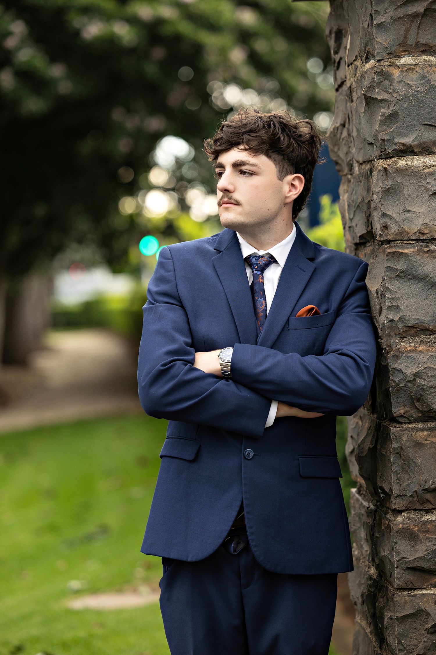 Young man in navy blue suit with tie and pocket square, arms crossed, leaning against stone pillar outdoors.