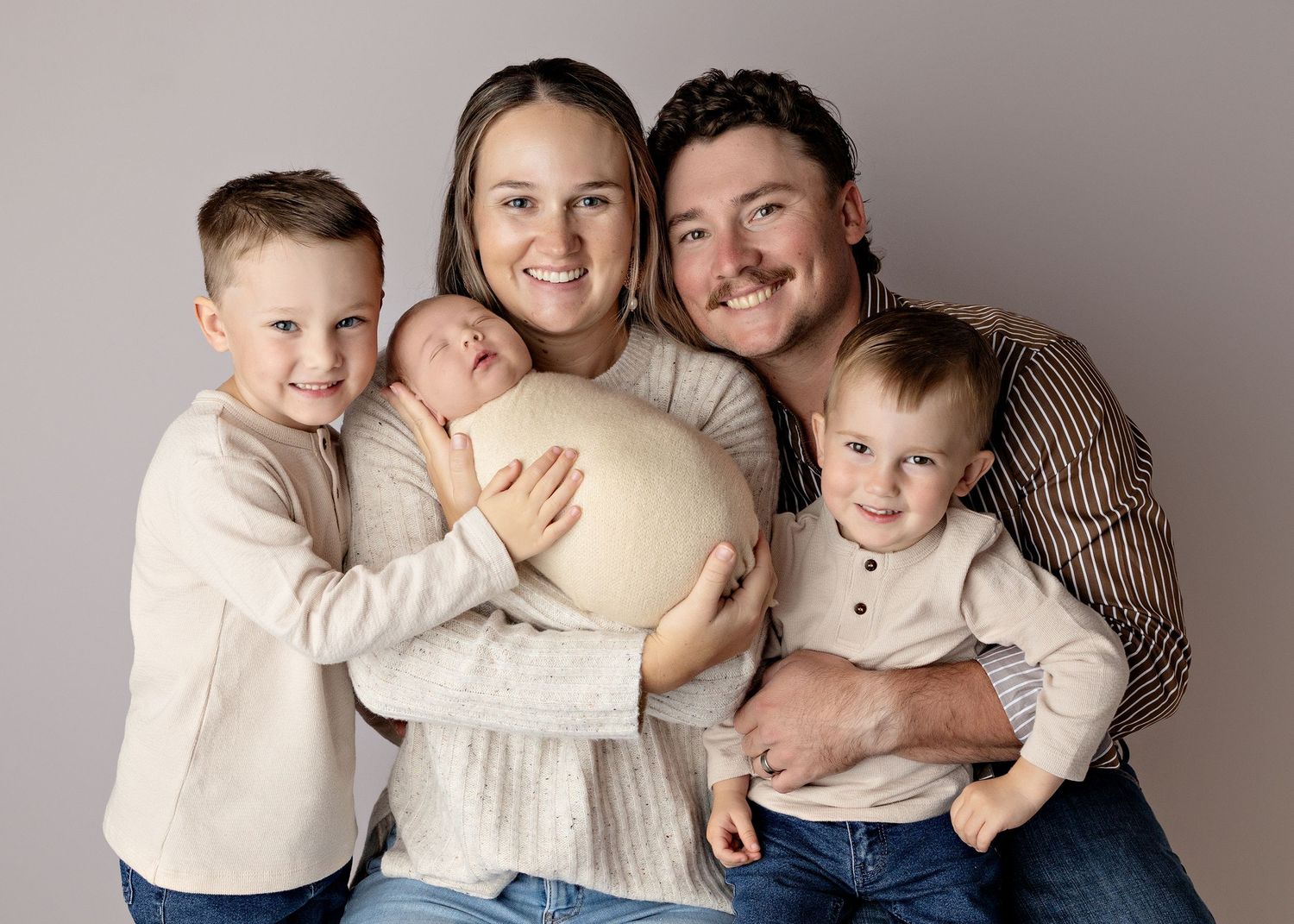 Happy family of five posing for a newborn portrait, parents with two young boys holding a swaddled baby.