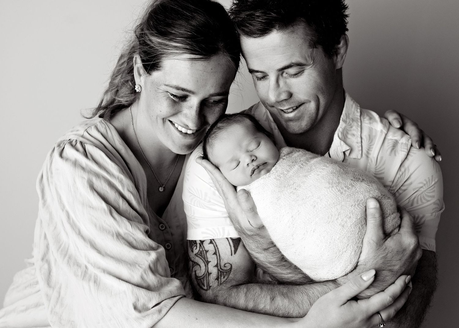 Black and white photo of smiling parents lovingly cradling their sleeping newborn baby together.