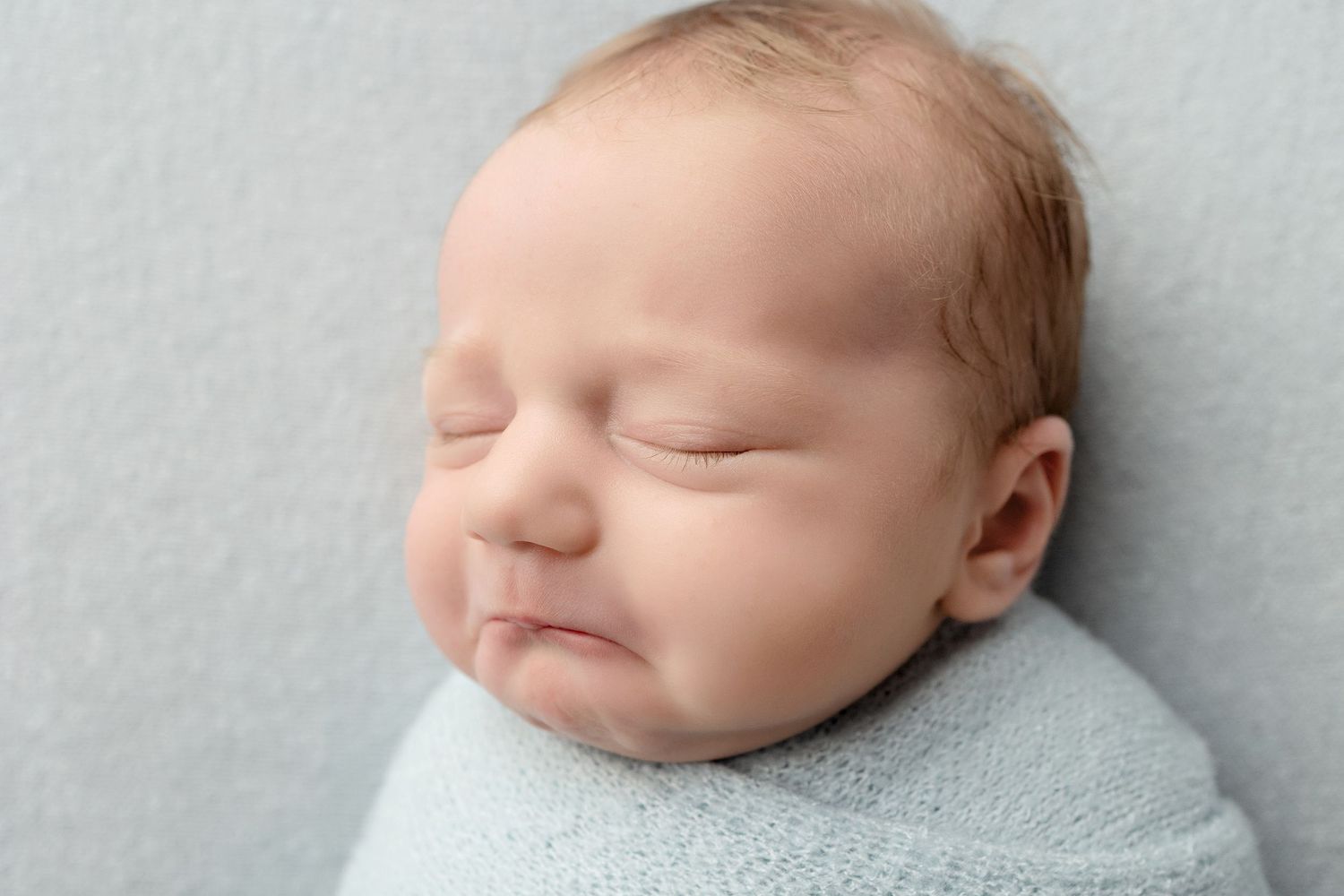 Peaceful sleeping newborn baby wrapped in soft light blue swaddle blanket on grey background.