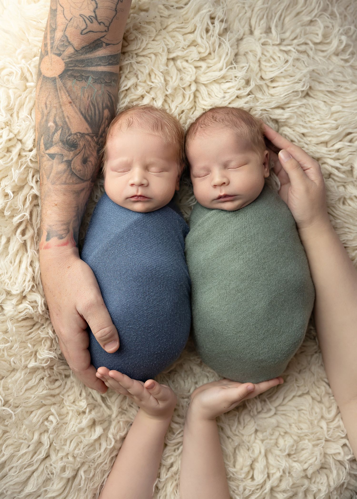 Two sleeping newborns in blue and green outfits lie side by side on a cream-colored fluffy rug while a tattooed hand touches them.Toowoomba