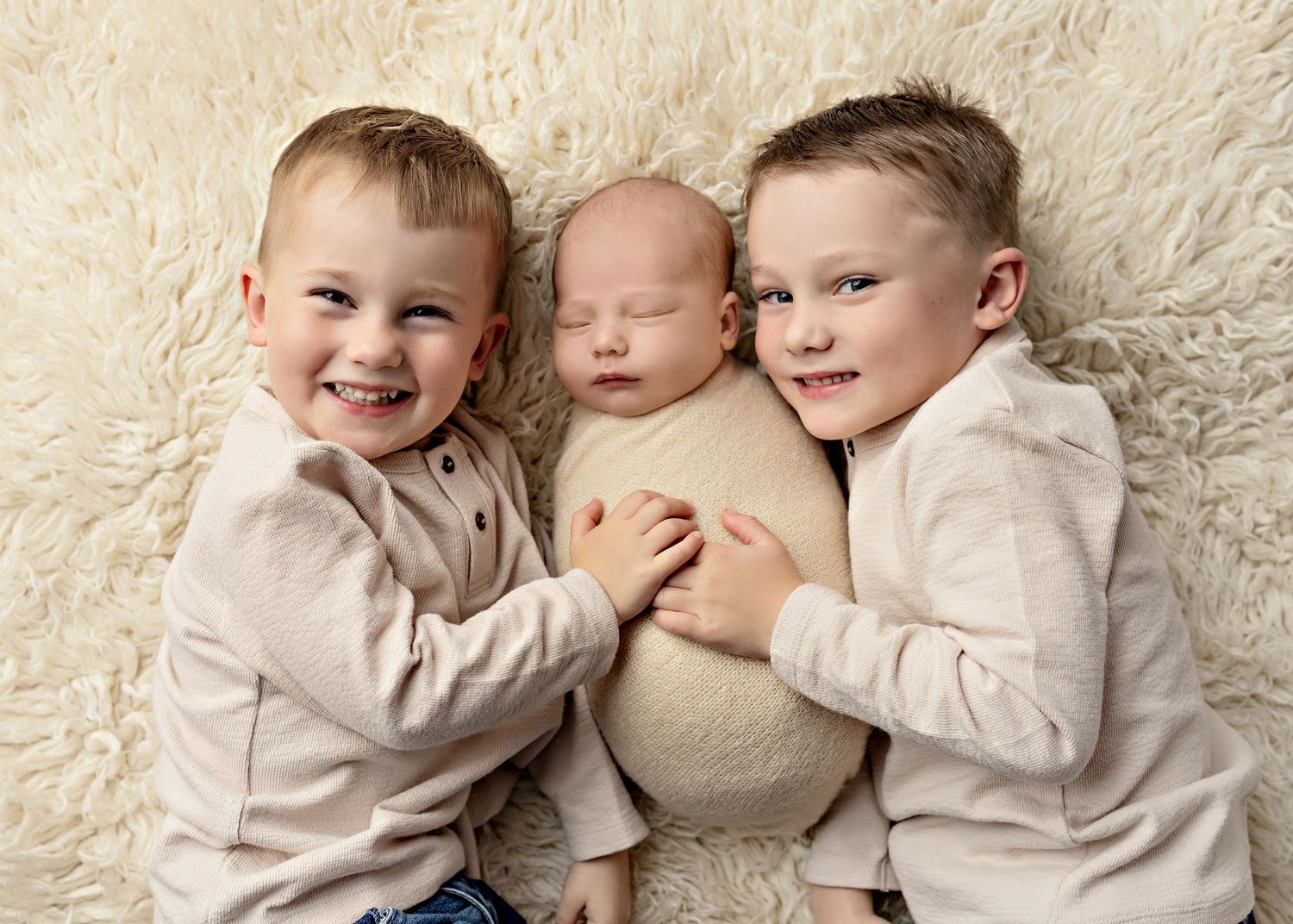 Two smiling toddler boys cuddle a sleeping newborn baby wrapped in cream, lying on a fluffy white rug.
