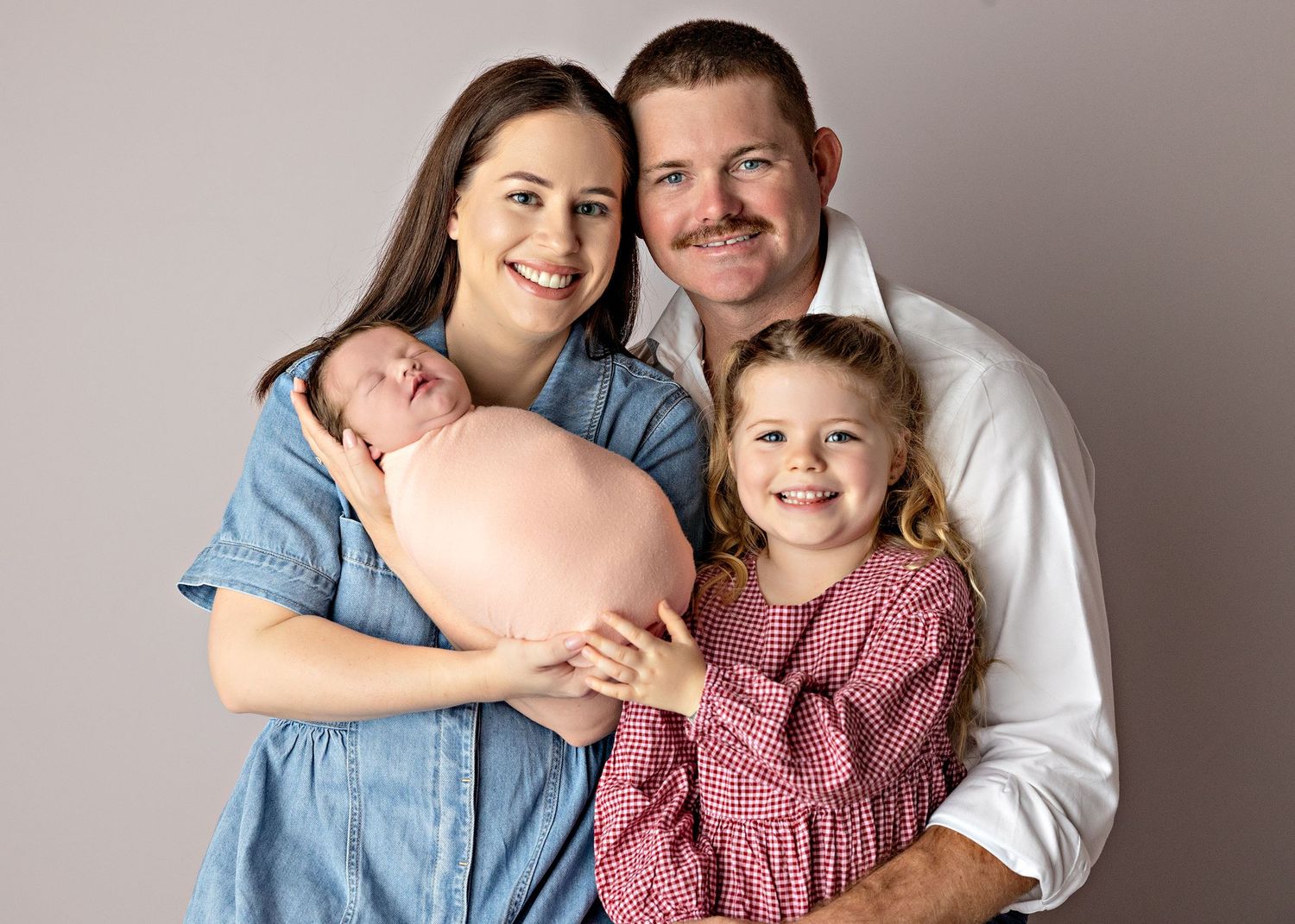 Family of four posing for newborn portrait, parents smiling with toddler girl and sleeping newborn baby in pink wrap.