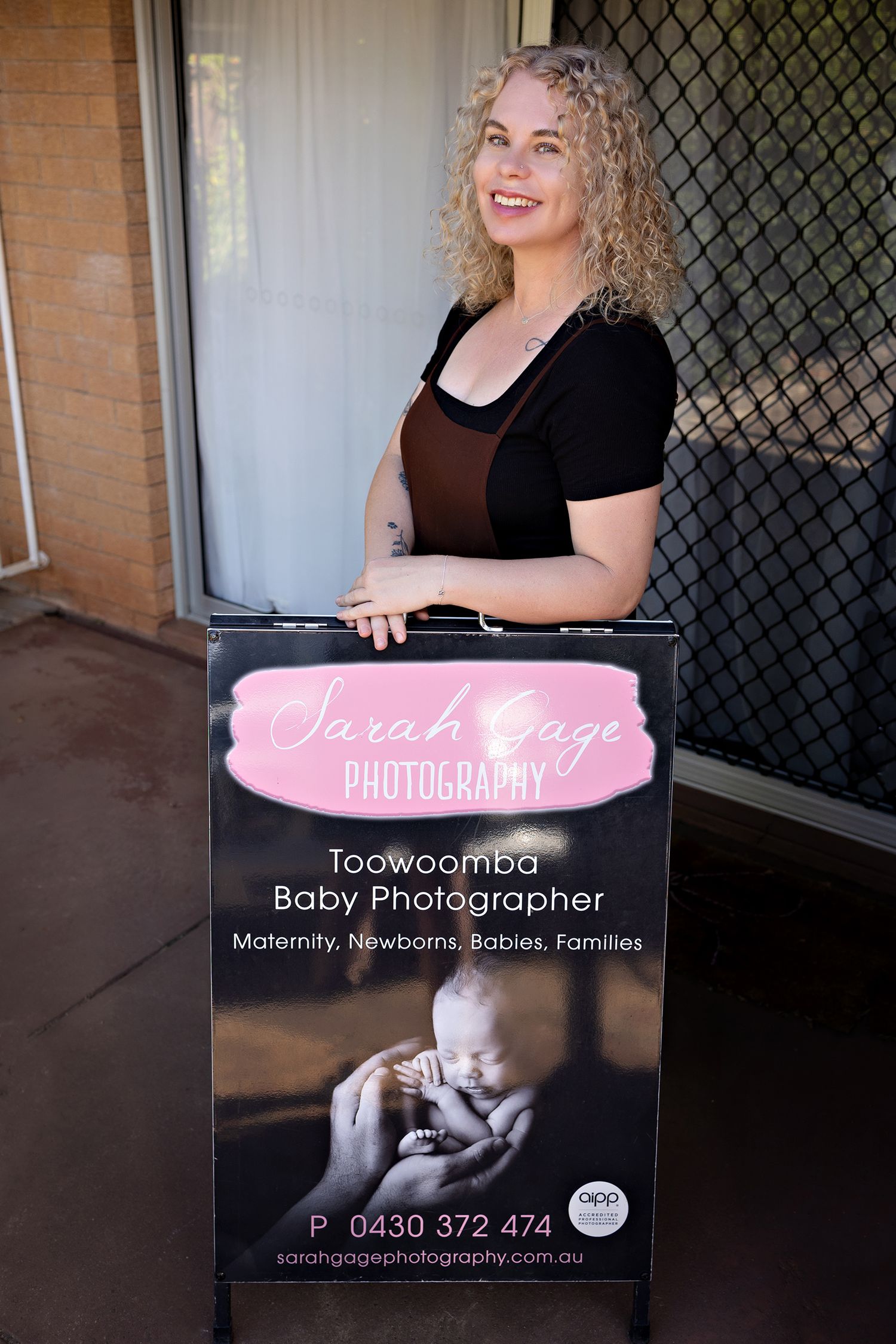 Professional photographer in black dress poses against chain link fence backdrop for business branding photos. Toowoomba Photographer