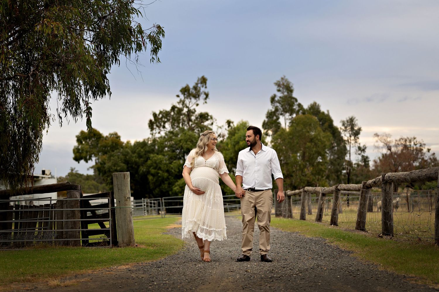 A couple walks down a tree-lined gravel path at sunset holding hands.