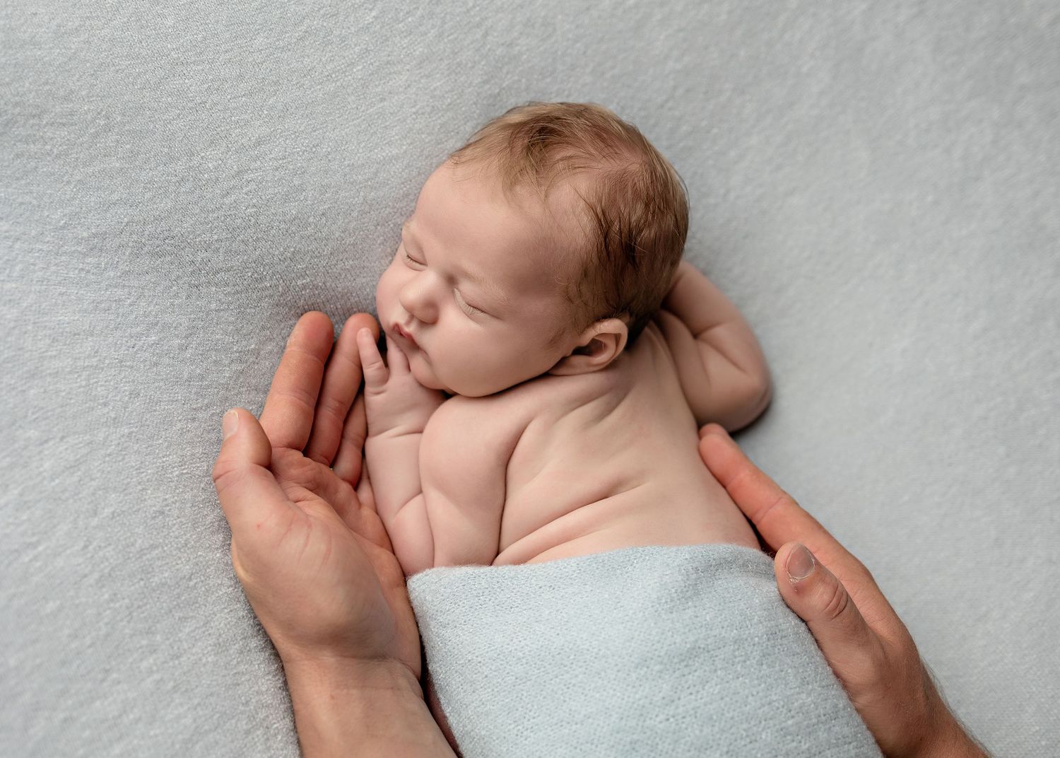 A newborn baby sleeps peacefully on white blanket while being cradled in adult hands.