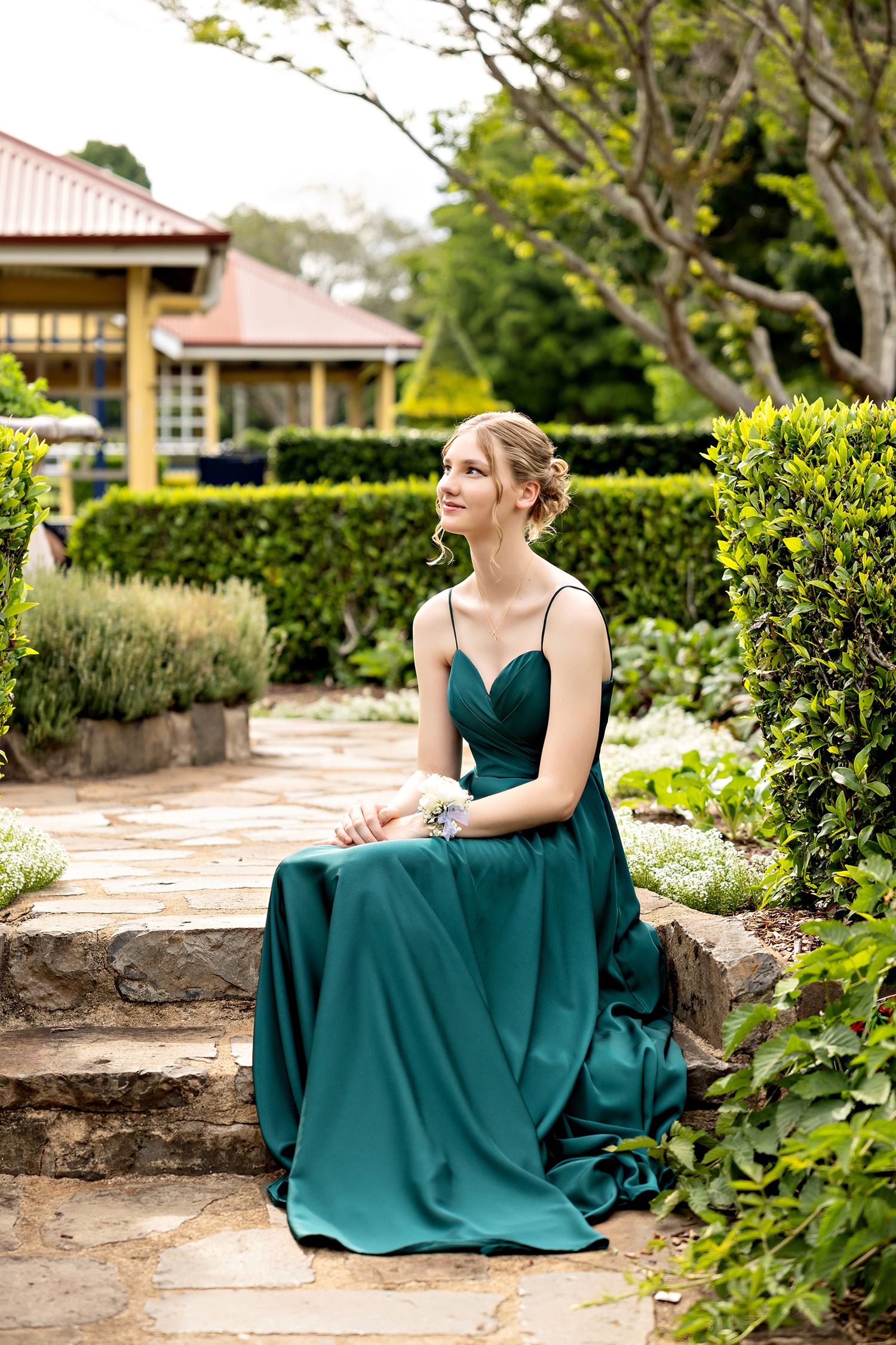 Person in emerald green formal gown sits elegantly on stone steps in garden setting.Toowoomba Photographer Toowoomba Newborn Photographer