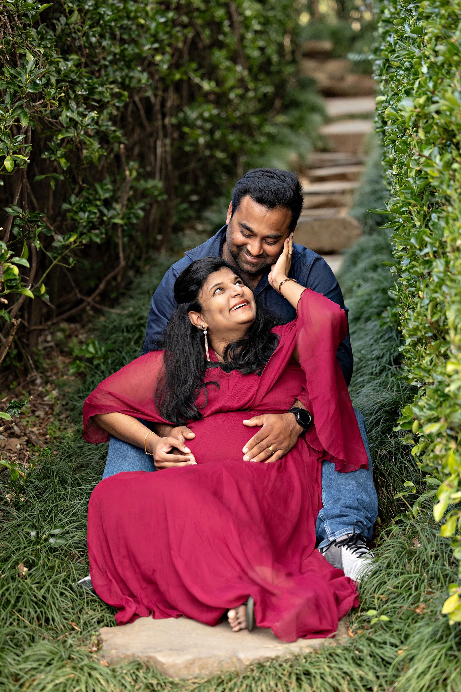 Couple shares intimate moment on garden steps surrounded by lush greenery, woman wearing flowing red dress.