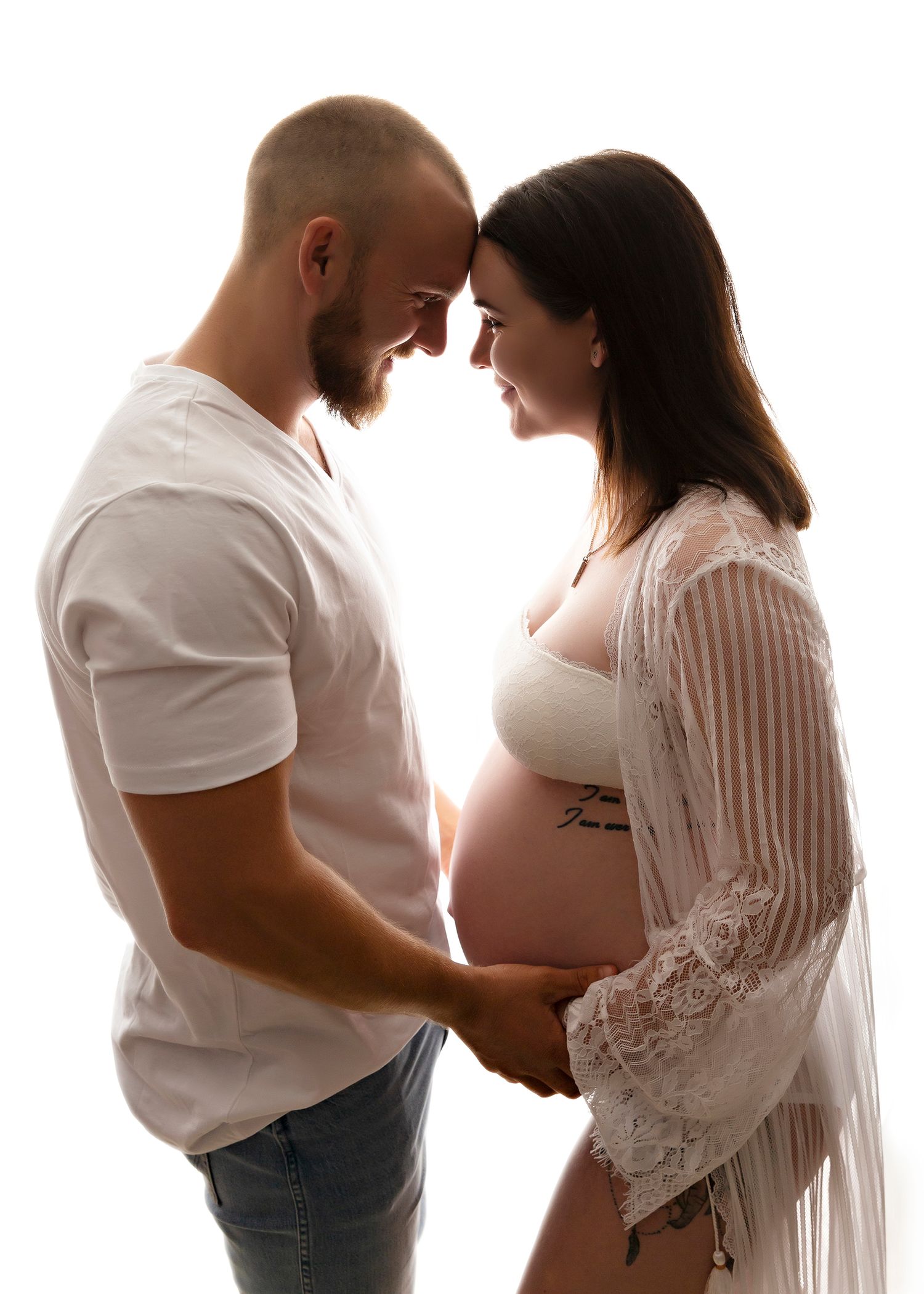 A romantic maternity photoshoot with a couple in white outfits against a bright background.