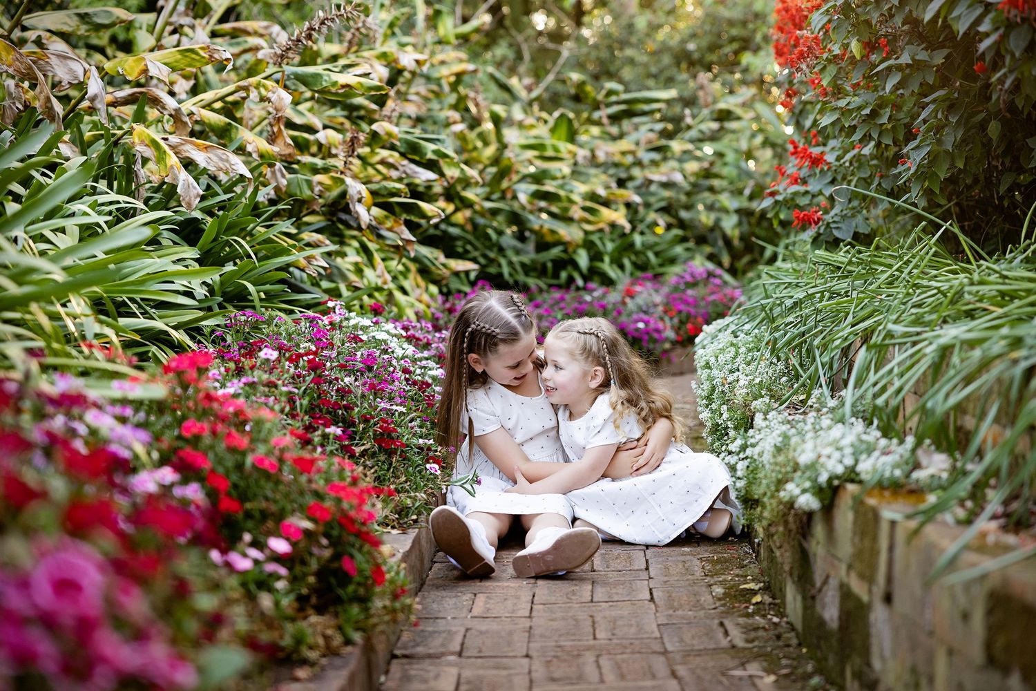 Two children sitting together on a brick pathway surrounded by colorful garden flowersToowoomba Photographer Toowoomba Newborn Photographer