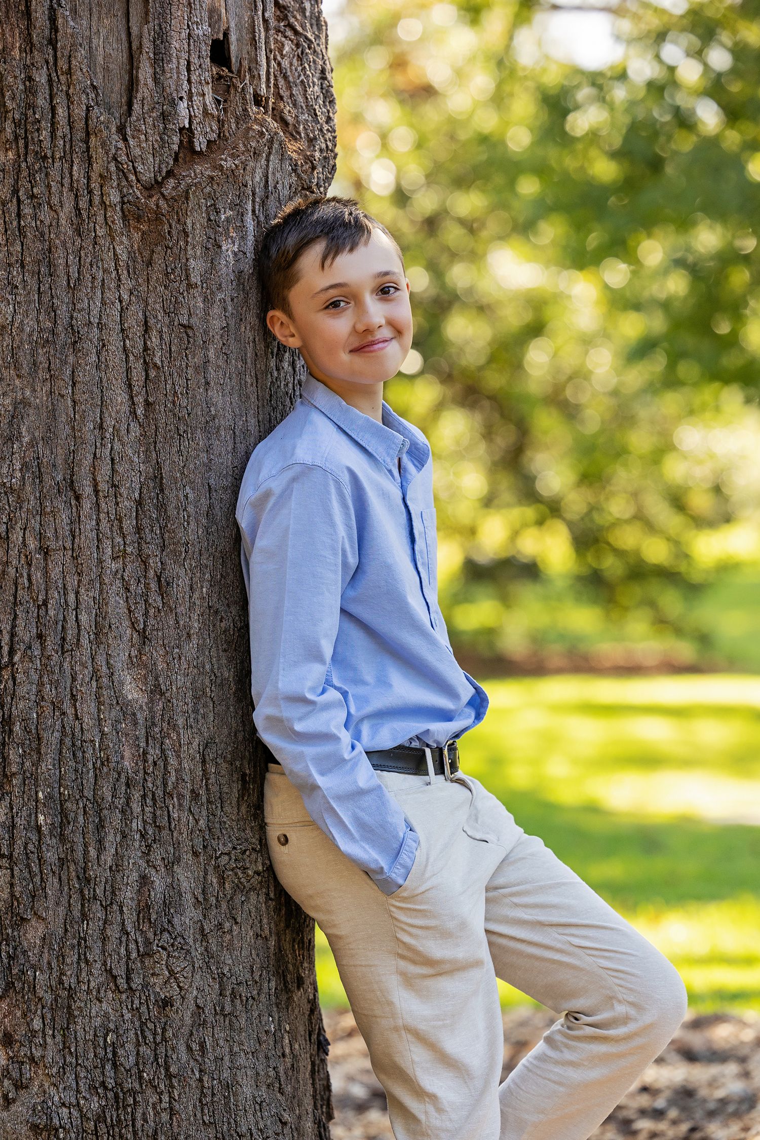 A smiling boy in a blue shirt and khaki pants leans against a large tree trunk in a sunny green park.
