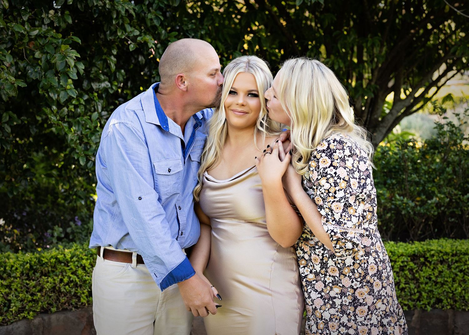 Three people share an intimate moment in a garden setting wearing formal attire.Toowoomba Photographer Toowoomba Newborn Photographer
