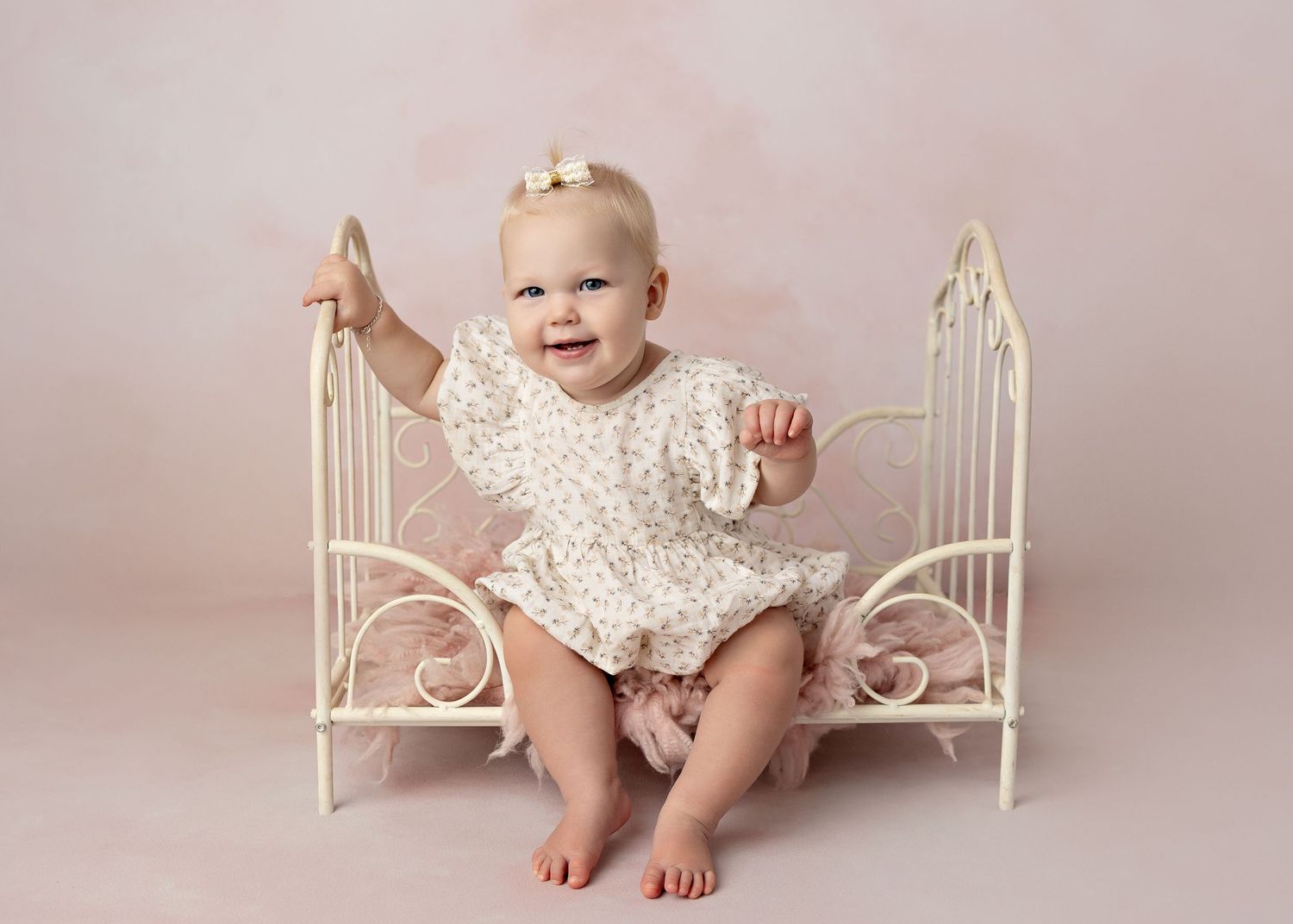 Smiling baby girl in floral romper sitting on white iron mini bed against pink backdrop.