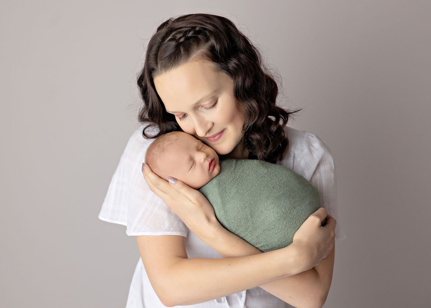 Parent in white shirt gently embraces sleeping newborn against neutral background.
