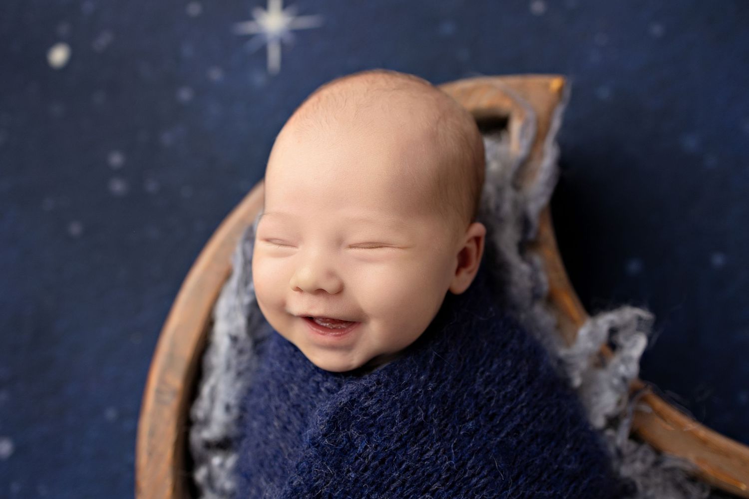 Smiling newborn baby wrapped in navy blue swaddle, sleeping in wooden bowl on starry night background.